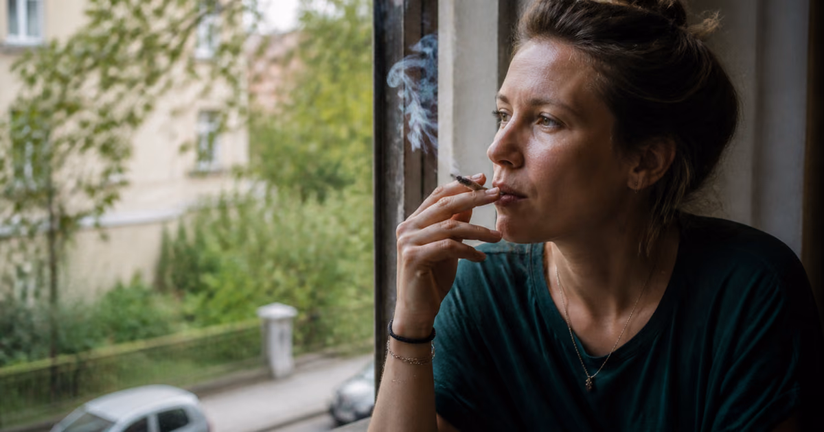 Жінка курить джойнт біля вікна, дивлячись на зелену вулицю